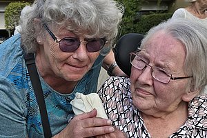 Zwei ältere Frauen sitzen nah beieinander, eine hält die Hand der anderen und beide tragen Brillen