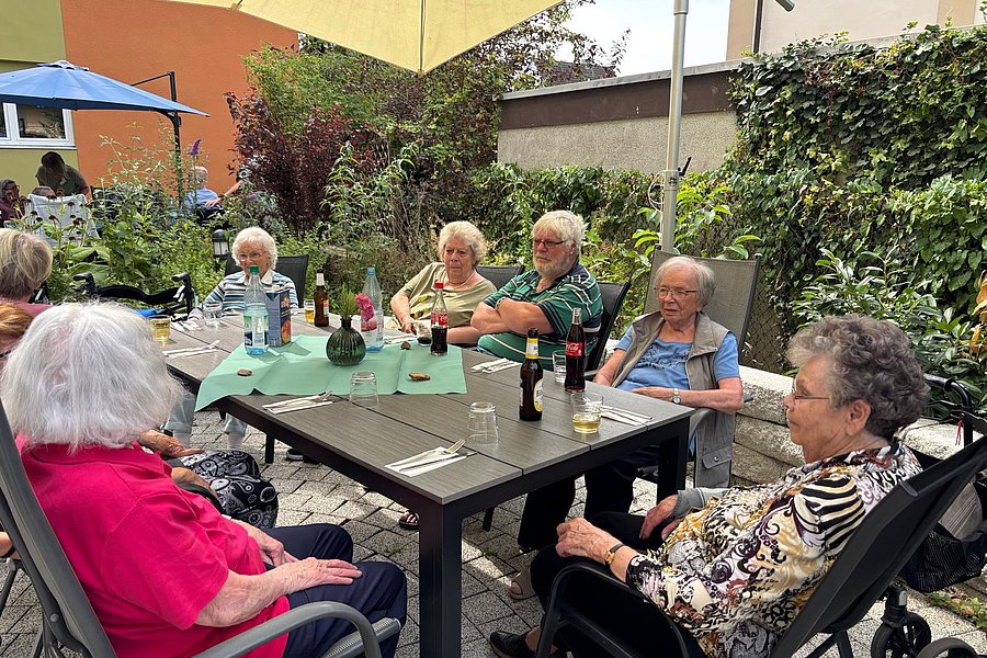 Mehrere ältere Menschen sitzen an einem rechteckigen Gartentisch unter einem Sonnenschirm im Freien und unterhalten sich
