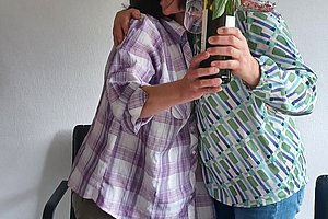 Zwei Frauen umarmen sich, eine hält eine Flasche Sekt mit Blumen, Freude und Feierlichkeit im Bild.