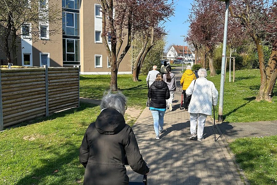 Gruppe älterer Menschen mit Jacken, die auf einem gepflasterten Weg in einem Wohngebiet spazieren gehen.