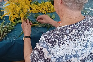 Zwei ältere Frauen sitzen an einem Tisch und binden Kränze aus gelben Blumen