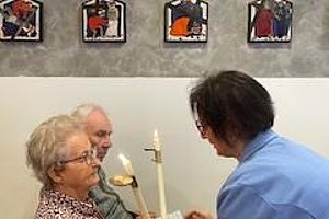 Zwei Senioren in einem Raum, eine Person zündet eine Kerze an, Bilder an der Wand im Hintergrund.