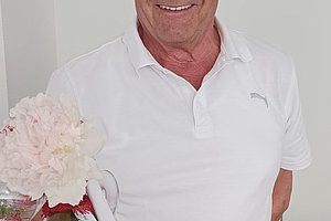 Lächelnder Mann in weißem T-Shirt hält eine Flasche Wein mit einer Blume in der Hand.