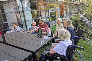Mehrere ältere Menschen sitzen an einem langen Gartentisch, eine Person im Rollstuhl, im Hintergrund ein modernes Gebäude mit großen Fenstern