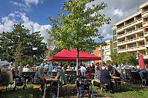 Menschen sitzen an Bänken und Tischen unter einem roten Pavillon im Freien neben einem Baum und Wohngebäuden
