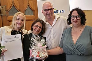 Vier lächelnde Personen stehen eng beieinander, eine Frau hält eine Urkunde, eine andere ein Geschenk mit Blumen, alle tragen festliche Kleidung.