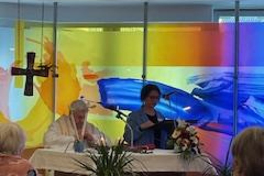Veranstaltung in einem Saal mit buntem Hintergrund, einer Person am Altar und mehreren Zuschauern im Hintergrund.