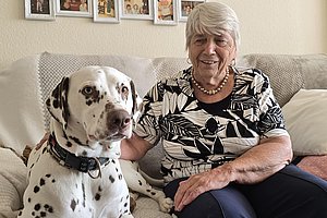 Ältere Frau mit kurzen weißen Haaren und Perlenkette sitzt auf einem Sofa und hält einen Dalmatiner mit Halsband an der Seite