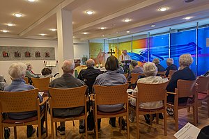 Gruppe älterer Menschen sitzt in einem Raum mit Holzboden auf Stühlen und hört einem Redner vor einer Wand mit großem farbigem Wandbild zu