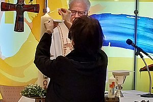 Person zündet in einer Kirche eine Kerze an, im Hintergrund ein Priester und ein Kreuz an der Wand
