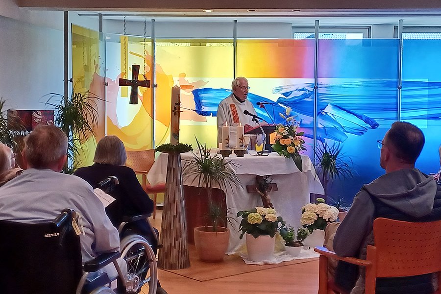 Ein Priester steht an einem mit Blumen geschmückten Altar in einem Raum mit mehreren sitzenden Menschen, einige in Rollstühlen, vor einer farbigen Glaswand mit abstraktem Muster.