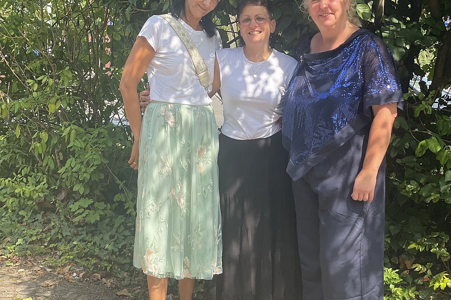Drei Frauen stehen nebeneinander auf einem gepflasterten Weg vor dichtem Grün.