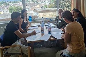 Fünf Personen sitzen an einem Tisch mit Papieren und Wasserflaschen vor einem großen Fenster mit Blick auf eine Stadtlandschaft