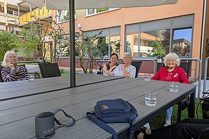 Mehrere Personen sitzen an einem großen Außentisch mit Getränkegläsern vor einem Wohngebäude
