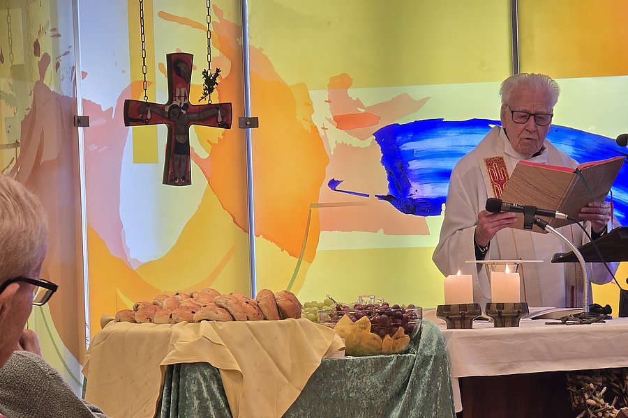 Priester mit Buch hinter Altar mit Kerzen und gedecktem Tisch mit Brot und Trauben in Kapelle mit farbiger Glaswand und hängendem Kreuz