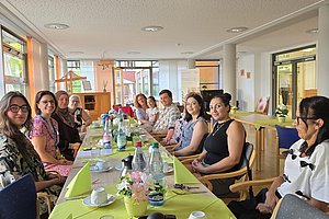 Gruppe von Menschen unterschiedlichen Alters sitzt an einem langen Tisch mit grünem Tischtuch in einem hellen Raum