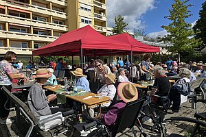 Gruppe älterer Menschen sitzt an Tischen unter rotem Pavillon im Freien vor Wohngebäude bei sonnigem Wetter