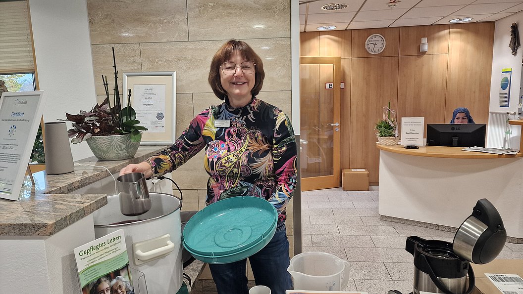 Frau mit buntem Shirt, die an einem Empfangstresen eine Kanne in eine grüne Schüssel schüttet.