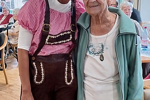 Zwei Personen in einem Raum mit Tischen und Stühlen, eine trägt eine Lederhose und ein rotes Hemd, die andere ein helles Oberteil und eine gemusterte Hose, im Hintergrund hängen bayerische Wimpelketten.