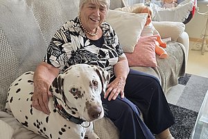 Ältere Frau mit buntem Oberteil sitzt auf einem Sofa neben ihrem süßen Dalmatiner-Hund.