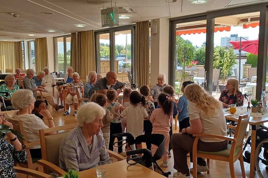 Mehrere ältere Menschen und Kinder sitzen in einem hellen Raum mit großen Fenstern an Tischen und unterhalten sich