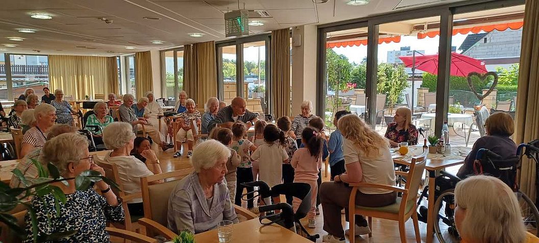 Mehrere ältere Menschen und Kinder sitzen in einem hellen Raum mit großen Fenstern an Tischen und unterhalten sich