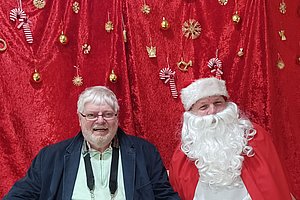 Weihnachtliche Szene mit zwei Männern, einer im Weihnachtsmann-Kostüm vor rotem Hintergrund mit Dekoration.