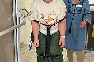 Eine ältere Frau mit weißer Haube, T-Shirt und grüner Hose geht mit einem schwarzen Gurt um die Taille, begleitet von einer lächelnden Frau in blauem Kleid und Brille in einem Flur.