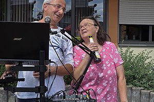 Zwei Personen stehen an einem Mikrofon und einem Laptop, die eine Person hält ein Mikrofon, die andere lächelt und lehnt sich leicht zur anderen Person