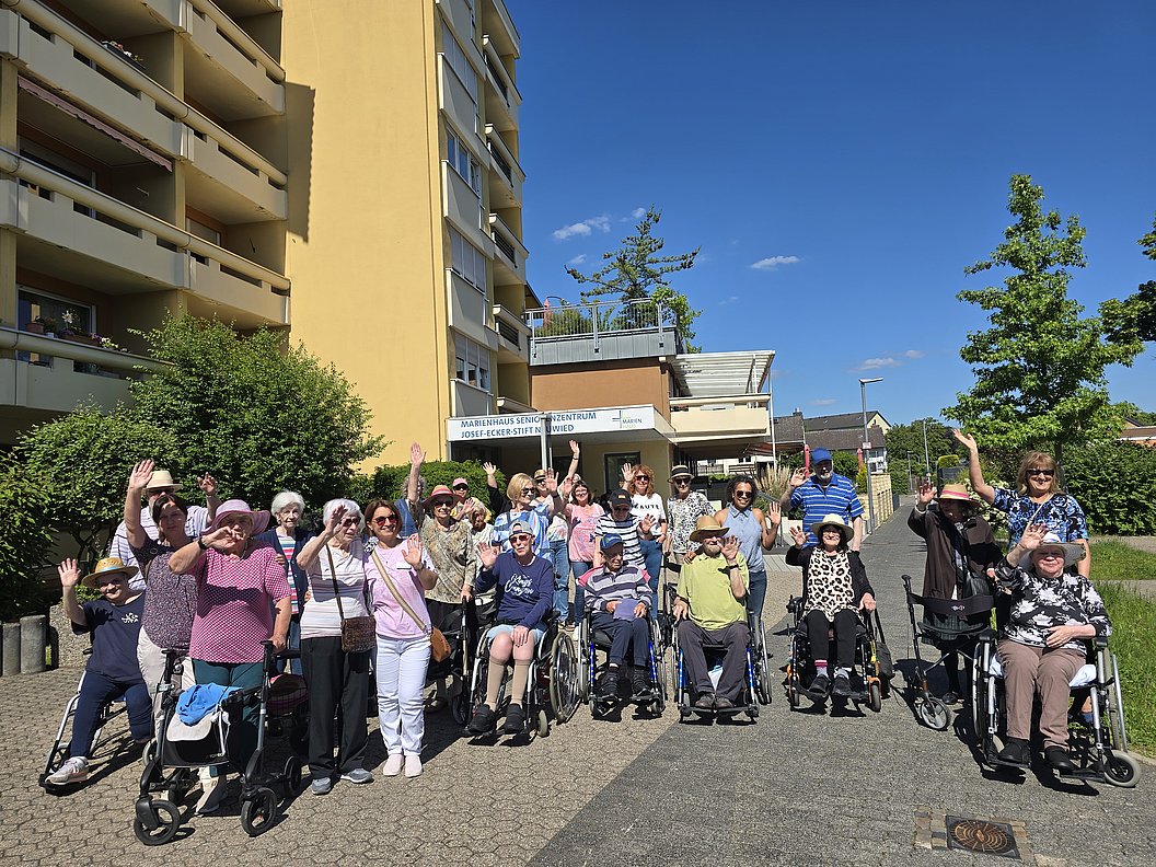Gruppe älterer Menschen im Freien, fröhlich winkend vor einem Wohnheim unter blauem Himmel.