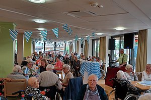 Großer Raum mit vielen älteren Menschen, die an Tischen sitzen und sich unterhalten, dekoriert mit blau-weiß karierten Wimpeln und Tischdecken, im Hintergrund steht eine Person an einem Rednerpult