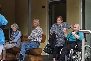 Vier ältere Menschen sitzen und stehen vor einem Gebäude, eine Frau sitzt im Rollstuhl und gestikuliert mit den Händen