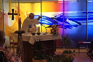 Priester hebt Kelch während einer Messe vor farbiger Glaswand mit abstraktem blau-gelbem Muster