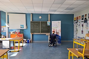 Klassenzimmer mit blauer Wand, Tafel mit Kreideschrift, mehreren Tischen und Stühlen sowie einer Person im Rollstuhl vor einem Informationsbanner
