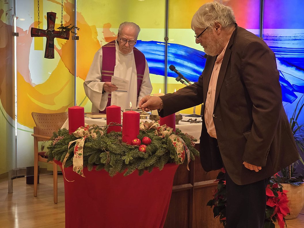 Zwei Männer stehen an einem Adventskranz mit roten Kerzen, eine von ihnen zündet eine Kerze an.