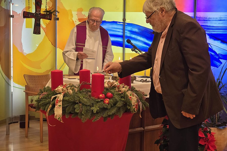 Zwei Männer stehen an einem Adventskranz mit roten Kerzen, eine von ihnen zündet eine Kerze an.