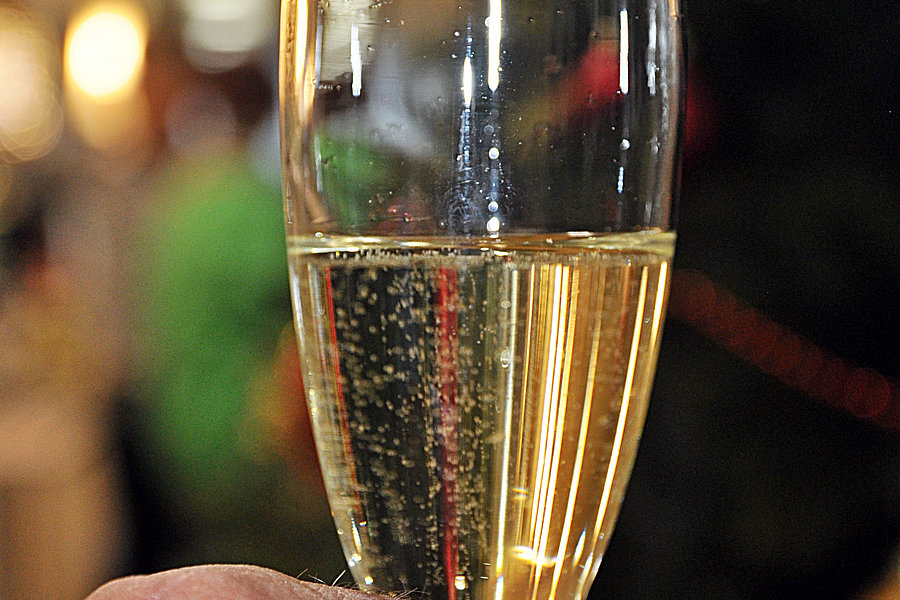 Hand mit Ehering hält ein Glas Sekt vor unscharfem Hintergrund mit Lichtern.