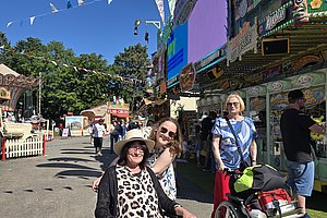 Zwei Frauen im Rollstuhl auf einem belebten Jahrmarkt mit Fahrgeschäften und bunten Fahnen im Hintergrund