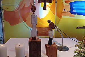 Holzfiguren auf einem Tisch mit Kerzen, Mikrofon und Blumenarrangement im Hintergrund.