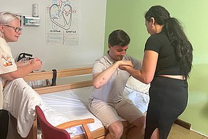 Mann sitzt auf Krankenhausbett und hält die Hand einer Frau, eine ältere Frau sitzt daneben auf einem Stuhl, im Hintergrund eine Zeichnung des Herzens an der Wand