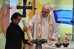 Zwei Personen in einer Kirche, eine hält eine große Kerze, im Hintergrund ein Kreuz und bunte Glasfenster