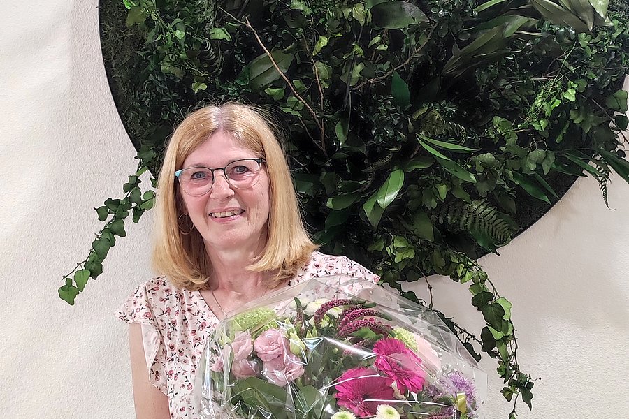 Frau steht vor einer Wand mit rundem, grünem Pflanzenarrangement und hält einen großen Blumenstrauß mit verschiedenen Blüten.