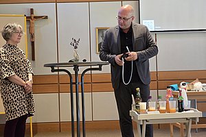 Präsentation in einem Saal: Mann mit Drinks hinter einem Tisch, Frau im Musterkleid beobachtet.