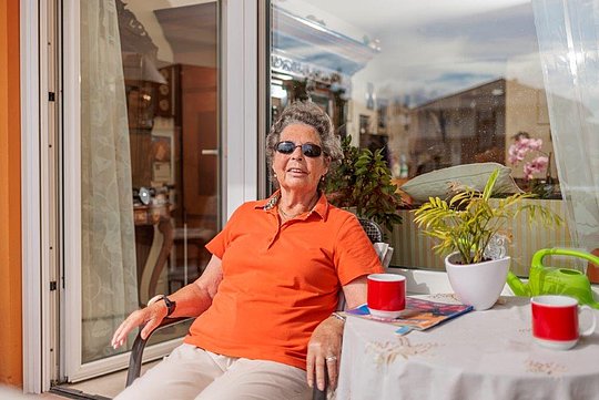 Ältere Frau in orangefarbenem Poloshirt sitzt auf Balkonstuhl neben Tisch mit zwei roten Tassen und Pflanzen.