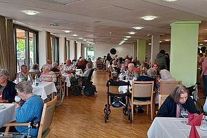 Großer Speisesaal mit mehreren Tischen, an denen ältere Menschen sitzen und sich unterhalten, einige mit Rollatoren und Gehhilfen
