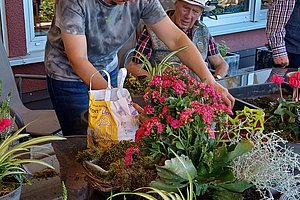Zwei Personen an einem Tisch mit verschiedenen Topfpflanzen und Blumenerde vor einem Fenster