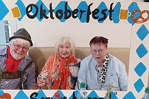 Drei Personen sitzen auf einer Bank hinter einem blau-weißen Oktoberfest-Rahmen mit Brezeln und Bierkrügen als Dekoration
