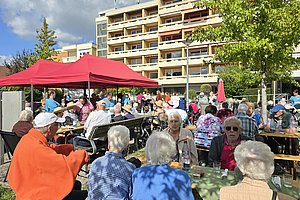 Ältere Menschen sitzen an Tischen im Freien unter einem roten Pavillon vor einem mehrstöckigen Wohngebäude an einem sonnigen Tag
