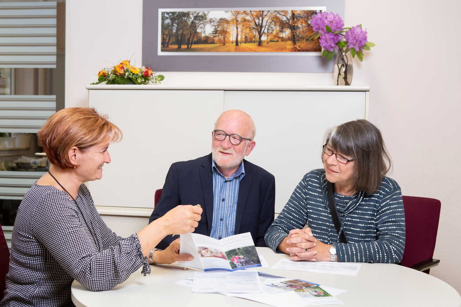 Drei Personen sitzen an einem runden Tisch mit geöffneten Broschüren, im Hintergrund ein weißer Schrank mit Blumen und ein Bild mit Herbstbäumen.