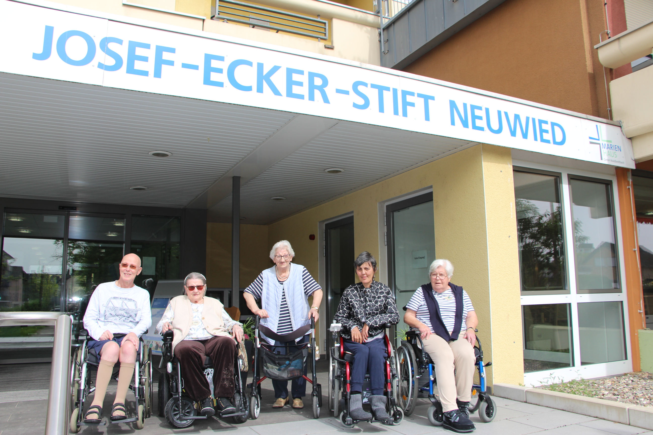 Fünf Personen, drei im Rollstuhl, vor dem Eingang des Josef-Ecker-Stifts Neuwied mit blau-weißer Beschriftung am Gebäude.